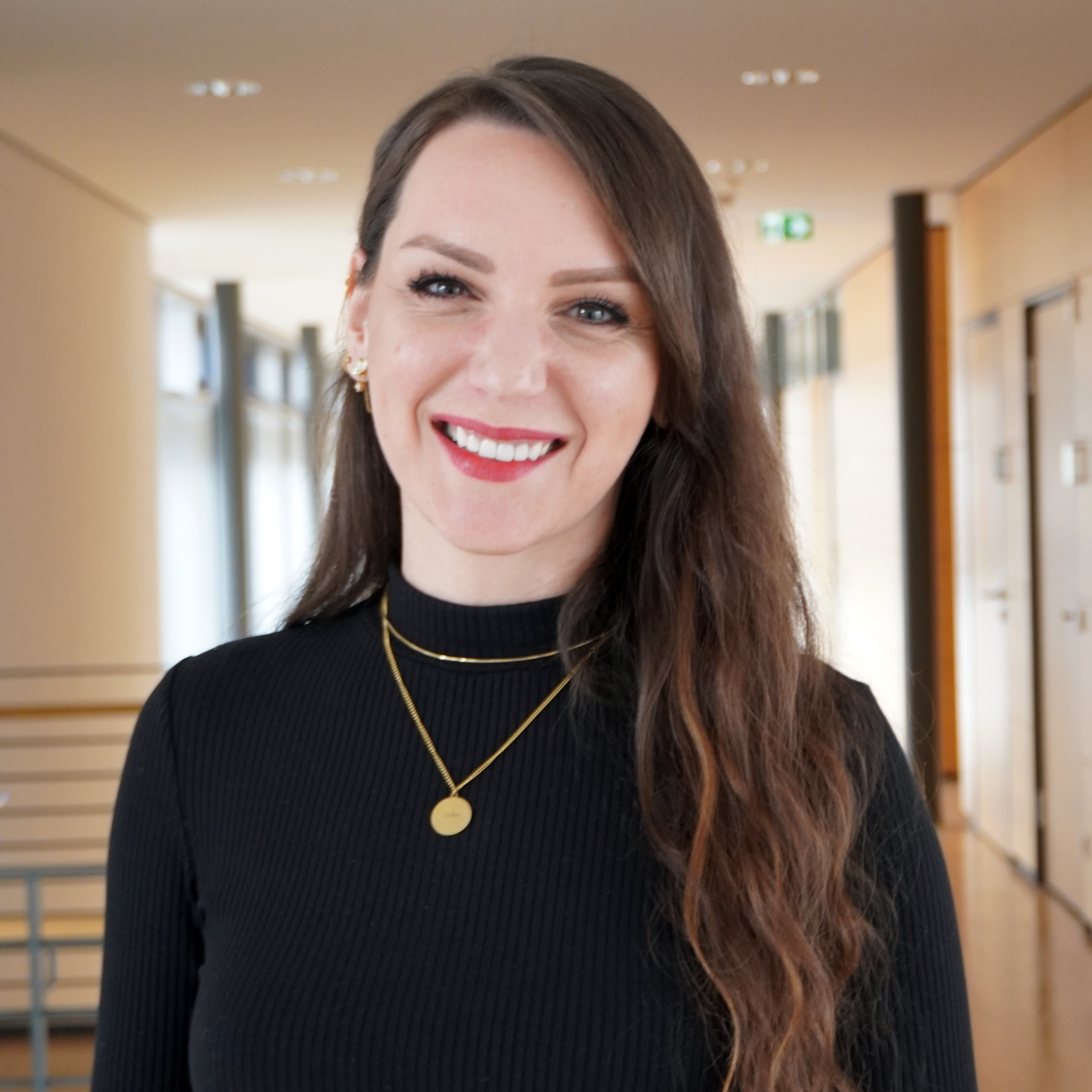 Une femme aux longs cheveux bouclés porte un haut noir et des bijoux. Elle sourit chaleureusement dans un intérieur moderne.