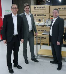 Quatre hommes en costumes se tiennent devant une table avec différents produits et des cartons de la marque HERMA en arrière-plan.