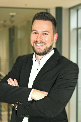 Homme en costume noir, les bras croisés, souriant amicalement dans un intérieur moderne.