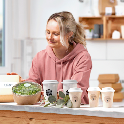 Une femme en sweat à capuche rose prépare des plats sains, dont un bol de salade et plusieurs tasses à café sur une table.