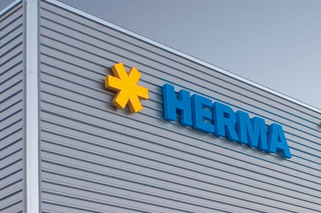Façade d'un bâtiment moderne avec le logo "HERMA" en lettres bleues et un symbole jaune.