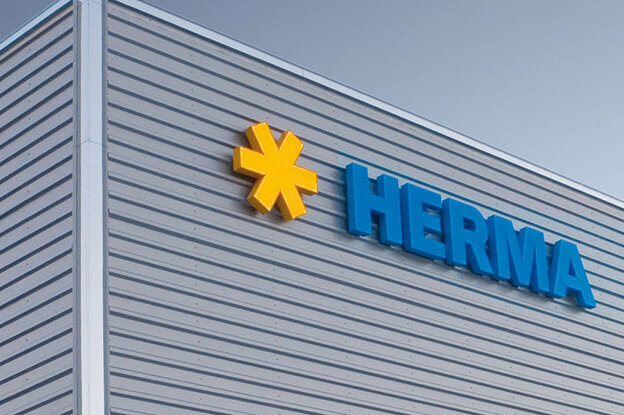 Façade d'un bâtiment moderne avec le logo "HERMA" en lettres bleues et un symbole jaune.