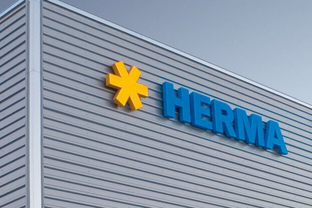 Façade d'un bâtiment moderne avec le logo "HERMA" en lettres bleues et un symbole jaune.