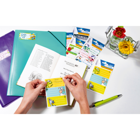 Hände halten eine gelbe Notizkarte mit einem Cartoon-Motiv, umgeben von Schulmaterialien, einem Buch und einer Vase mit Blumen.
