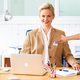 Une femme aux cheveux blonds est assise à un bureau, portant un blazer bleu et souriant. Sur la table se trouvent un ordinateur portable et du matériel d'écriture.