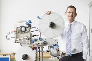 Un homme en chemise et cravate se tient à côté d'une machine technique avec des rouleaux et des éléments de commande.