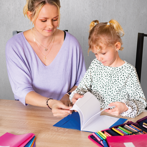 Ein Kind und ein Jugendlicher sitzen an einem Schreibtisch und malen. Im Hintergrund steht ein Regal mit Spielzeug und Büchern.