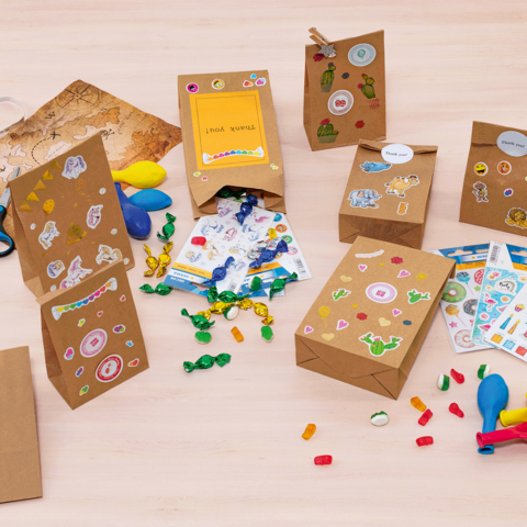 Des sacs-cadeaux colorés et du matériel de bricolage sur une table, comprenant des autocollants, des ballons, des ciseaux et des bonbons.