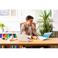 Junger Mann sitzt an einem Schreibtisch, arbeitet mit einem Laptop und einem Ordner, umgeben von Pflanzen und bunten Aktenordnern.