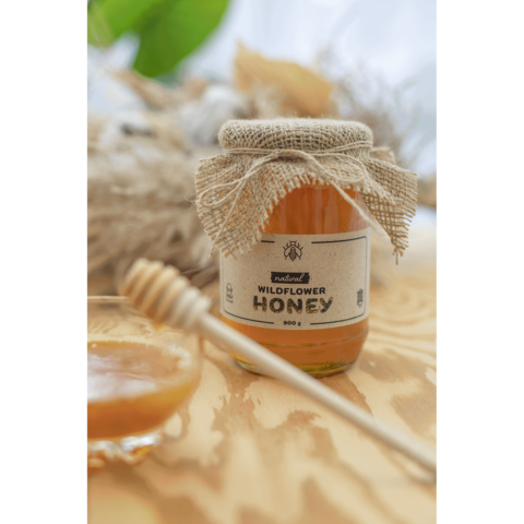 Glas Honig mit Jute-Deckel und Etikett "Wildflower Honey", daneben ein Holzlöffel und eine kleine Schale mit Honig.