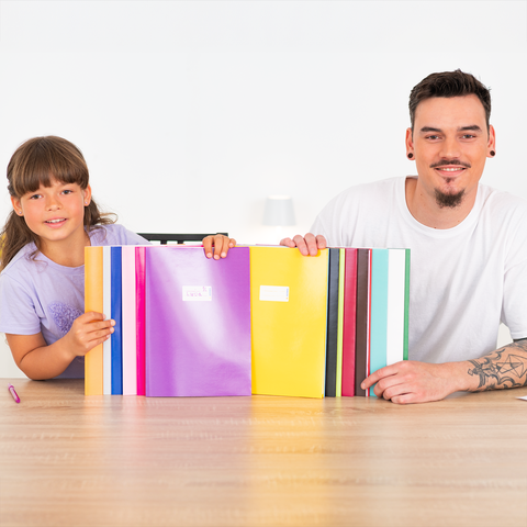 Ein Mädchen und ein Mann sitzen an einem Tisch, lächelnd, mit bunten Ordnern vor sich. Der Mann hat Tattoos und trägt ein weißes T-Shirt.
