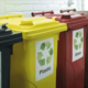 Drei Mülltonnen in verschiedenen Farben: gelb für Plastik, rot für Glas und schwarz für Restmüll, mit Recycling-Symbolen.