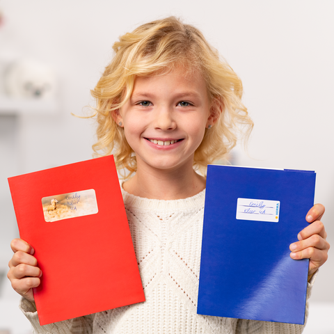 Un enfant souriant tient un cahier rouge et un cahier bleu dans les mains, tous deux ornés d'autocollants.