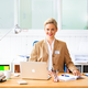 Une femme aux cheveux blonds est assise à un bureau, sourit et porte un blazer beige. Devant elle se trouvent un ordinateur portable et du matériel d'écriture.