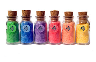 Six petites bouteilles en verre avec des bouchons, remplies de sable coloré dans les teintes vert, bleu, violet, rose, orange et jaune.