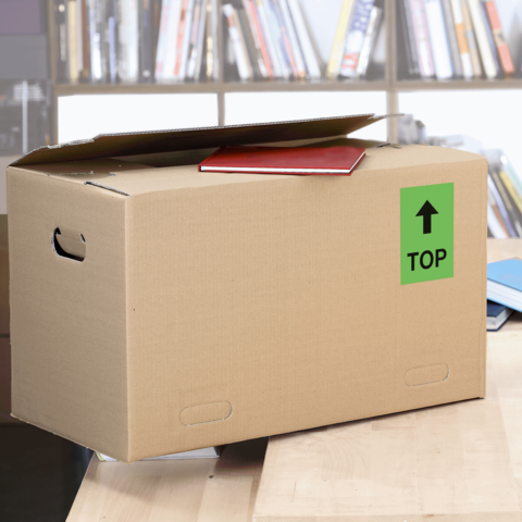 Kartonbox auf einem Tisch, mit einem roten Heft oben und einem grünen Aufkleber mit der Aufschrift "TOP".