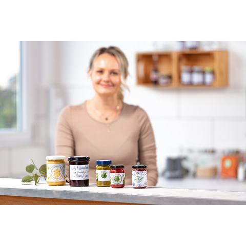 Eine Frau mit blonden Haaren steht an einer Küchentheke, umgeben von verschiedenen Marmeladengläsern.