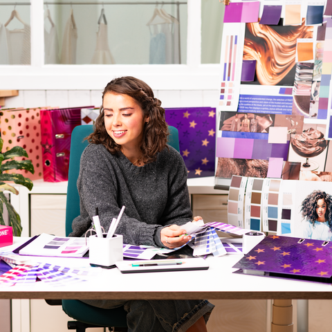 Une femme aux cheveux bouclés est assise à une table, entourée d'échantillons de couleurs et de matériaux de design, tandis qu'elle travaille sur un projet.