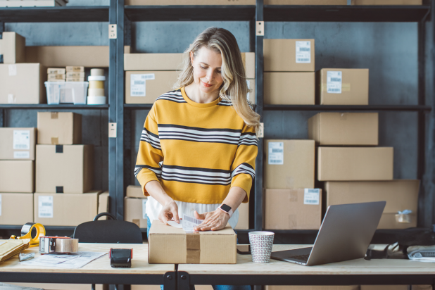 Une femme portant un pull rayé se tient à une table, entourée de cartons et d'un ordinateur portable dans un bureau moderne.