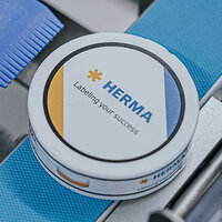 Boîte ronde avec l'impression "HERMA" et le slogan "Labeling your success", entourée d'éléments bleus et blancs.
