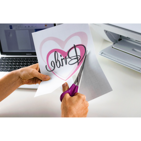 Eine Person schneidet mit einer Schere ein Blatt Papier mit einem Herz und dem Wort "Bride" aus, während ein Laptop im Hintergrund sichtbar ist.