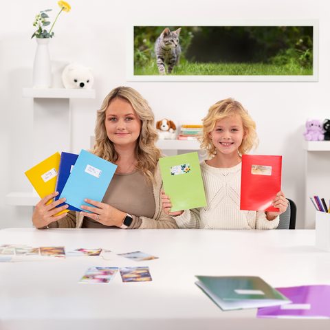 Une femme et une fille tiennent des classeurs colorés dans les mains, tandis qu'elles sont assises à une table avec des photos. En arrière-plan, on peut voir un chat.