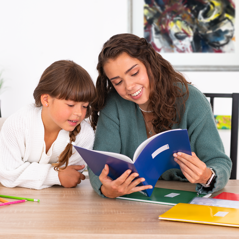 Eine junge Frau und ein Mädchen sitzen an einem Tisch und lesen gemeinsam in einem Buch, umgeben von bunten Heften und Stiften.