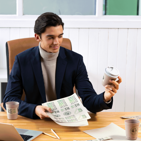 Ein junger Mann in einem Anzug sitzt an einem Tisch, hält einen Kaffeebecher und betrachtet Papiere mit Druckdesigns.