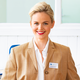 Une femme souriante aux cheveux blonds porte un blazer beige et une blouse blanche. Un badge avec le nom "Manuela Richter" est visible.