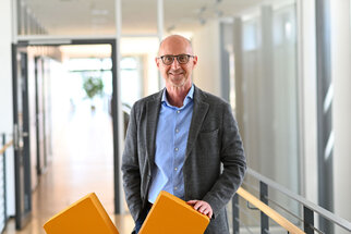 Un homme avec des lunettes se tient dans un couloir de bureau moderne et tient deux coussins orange. Il porte un blazer gris et une chemise bleue.