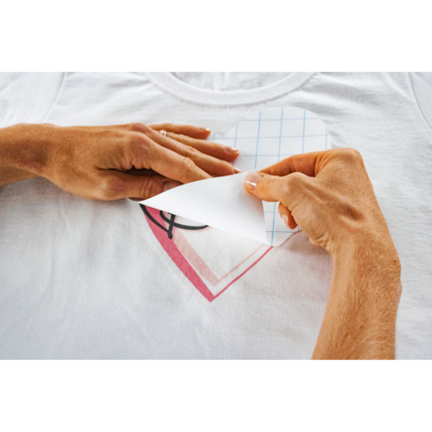 Hände entfernen eine Schutzfolie von einem bedruckten Herzmotiv auf einem weißen T-Shirt.