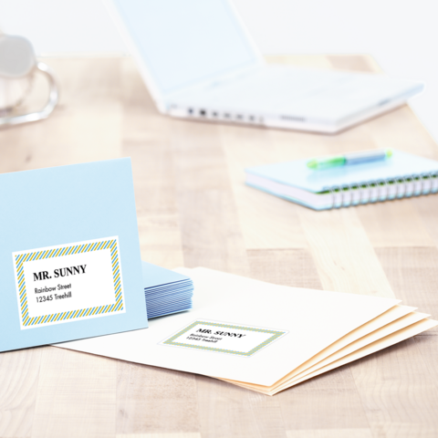 Des enveloppes bleues et blanches avec des étiquettes d'adresse sont posées sur une table, à côté d'un ordinateur portable et d'un carnet.