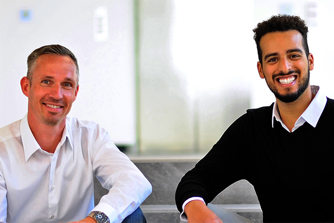 Deux hommes sont assis côte à côte sur une marche, souriant et portant des vêtements formels. L'arrière-plan est lumineux et moderne.