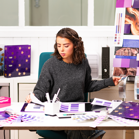 Une personne est assise à une table, entourée d'échantillons de couleurs et de matériaux de design, tout en travaillant sur un projet créatif.