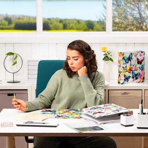 Une jeune femme est assise à un bureau, entourée de matériaux colorés et de notes, tout en tenant un stylo d'un air pensif.