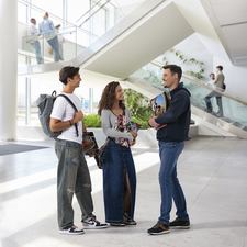 Drei junge Erwachsene stehen in einem modernen Raum und unterhalten sich. Sie tragen lässige Kleidung und halten Taschen. Im Hintergrund sind weitere Personen zu sehen.