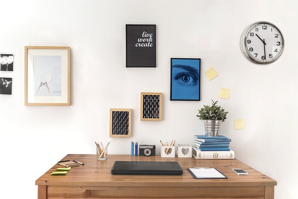 Un bureau avec un ordinateur portable, des images décoratives au mur, une plante et une lampe sur pied. Une horloge murale indique l'heure.