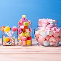Des bonbons colorés dans des bocaux, comprenant des sucettes, des oursons en gélatine et des guimauves en forme de cœur, sur un fond en bois clair.