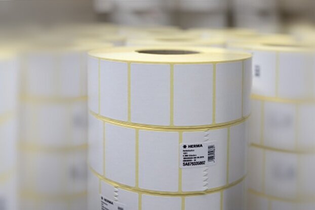 Rouleaux avec des étiquettes blanches, empilés dans un environnement de stockage. Les étiquettes sont marquées de lignes jaunes.