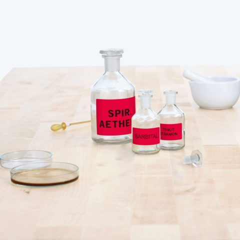 Flaschen mit roten Etiketten, beschriftet mit "Spir. Aether", "Barbital" und "Tinct. Tramon", sowie ein Mörser und zwei Glasplatten auf einem Tisch.
