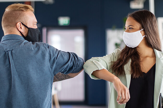 Deux personnes dans un bureau moderne se saluent en se touchant le coude, toutes deux portent des masques.
