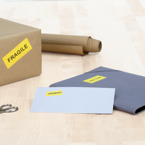 Ein Karton mit dem Aufkleber "FRAGILE", daneben ein graues Kuvert mit dem Aufkleber "URGENT!" und eine Schere auf einem Holztisch.