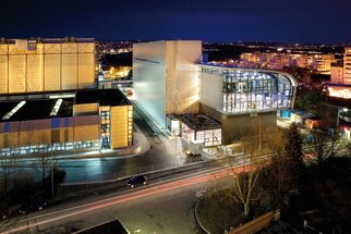 Architecture moderne d'un bâtiment la nuit, entouré de lumières urbaines et d'une rue illuminée par les phares des voitures.