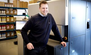 Un homme sourit à côté d'une presse dans un environnement de travail moderne, entouré d'étagères remplies de matériel d'impression.