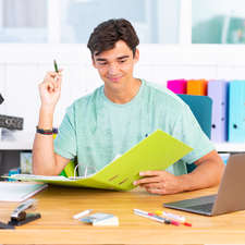 Ein junger Mann sitzt an einem Schreibtisch, hält einen Stift in der Hand und schaut auf einen offenen Ordner. Ein Laptop und Schreibmaterial sind sichtbar.