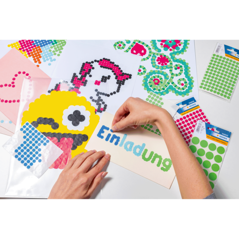 Hände gestalten Einladungen mit bunten Punkten auf Papier, darunter ein gelbes Monster und verschiedene Aufkleber in grünen und blauen Farben.