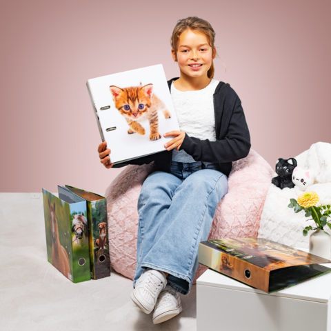 Un fille est assise sur un pouf et tient un classeur avec un chaton sur la couverture. Autour d'elle, il y a d'autres classeurs et une plante.