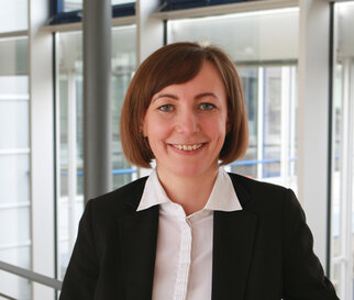 Une femme souriante avec des cheveux bruns mi-longs porte un blazer noir et une chemise blanche. Elle se tient dans un bâtiment de bureau moderne.