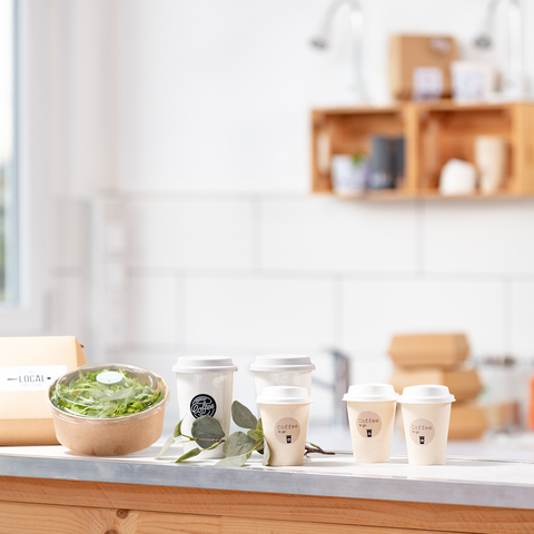 Une table en bois avec un saladier, plusieurs tasses à café et un emballage, entourée d'une cuisine lumineuse et moderne.