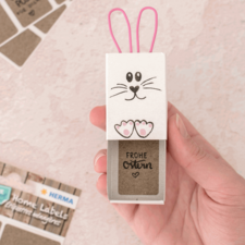 Eine Hand hält eine Verpackung in Form eines Hasen mit Ohren und einem Gesicht, die den Text "FROHE Ostern" zeigt.